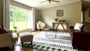 Una sala de estar con ventilador de techo, un ventanal con cortinas, un sofá color beige, un sillón blanco, una mesa de centro y un tapete estampado.
