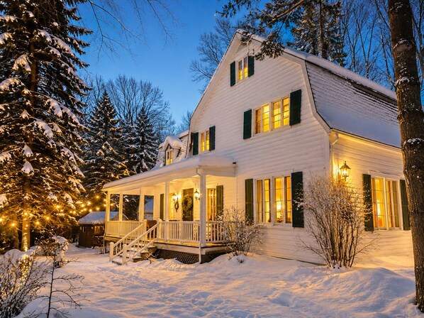 A two-story house with a porch and a covered entryway, surrounded by snow and trees.