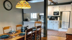 Un comedor con una mesa de madera dispuesta para cuatro personas, rodeada de sillas. Un reloj está colgado en la pared sobre la mesa.