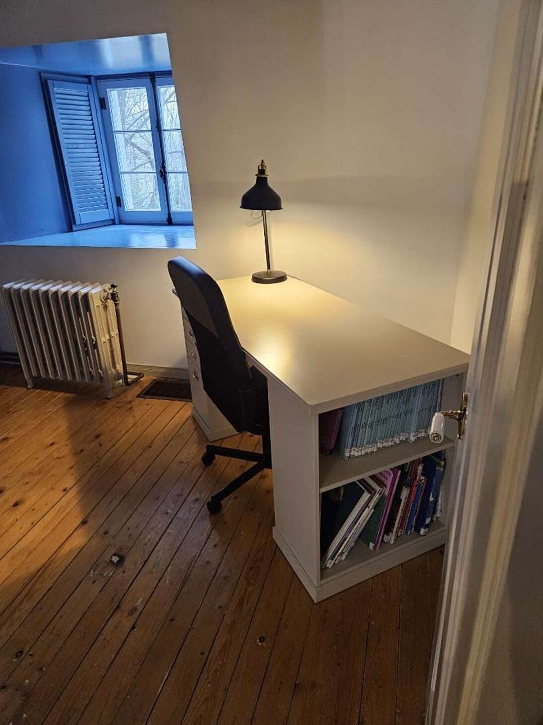 A small office space with a white desk, a black chair, a radiator, and a bookshelf with books.