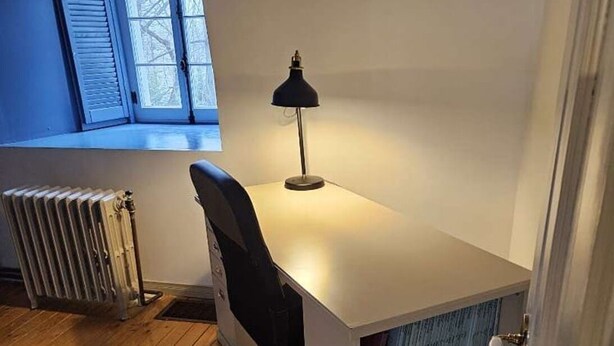 Un petit bureau avec un bureau blanc, une chaise noire, un radiateur et une bibliothèque remplie de livres.