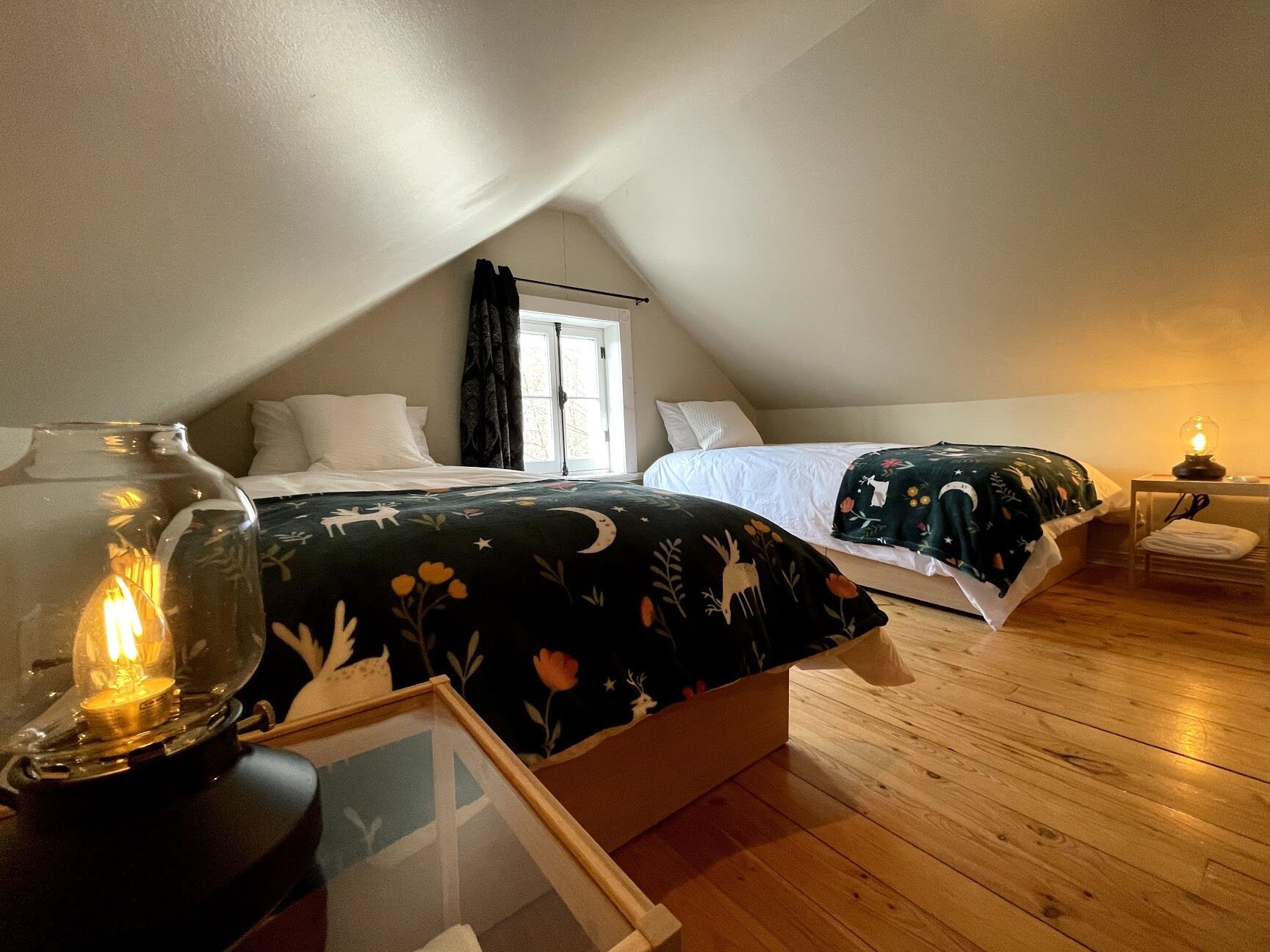 A cozy attic bedroom with two beds, a wooden floor, a window with curtains, and a table lamp.