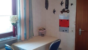 A small dining area with a table and two chairs, a window with curtains, a vase with flowers, and wall decorations including a compass and birds.