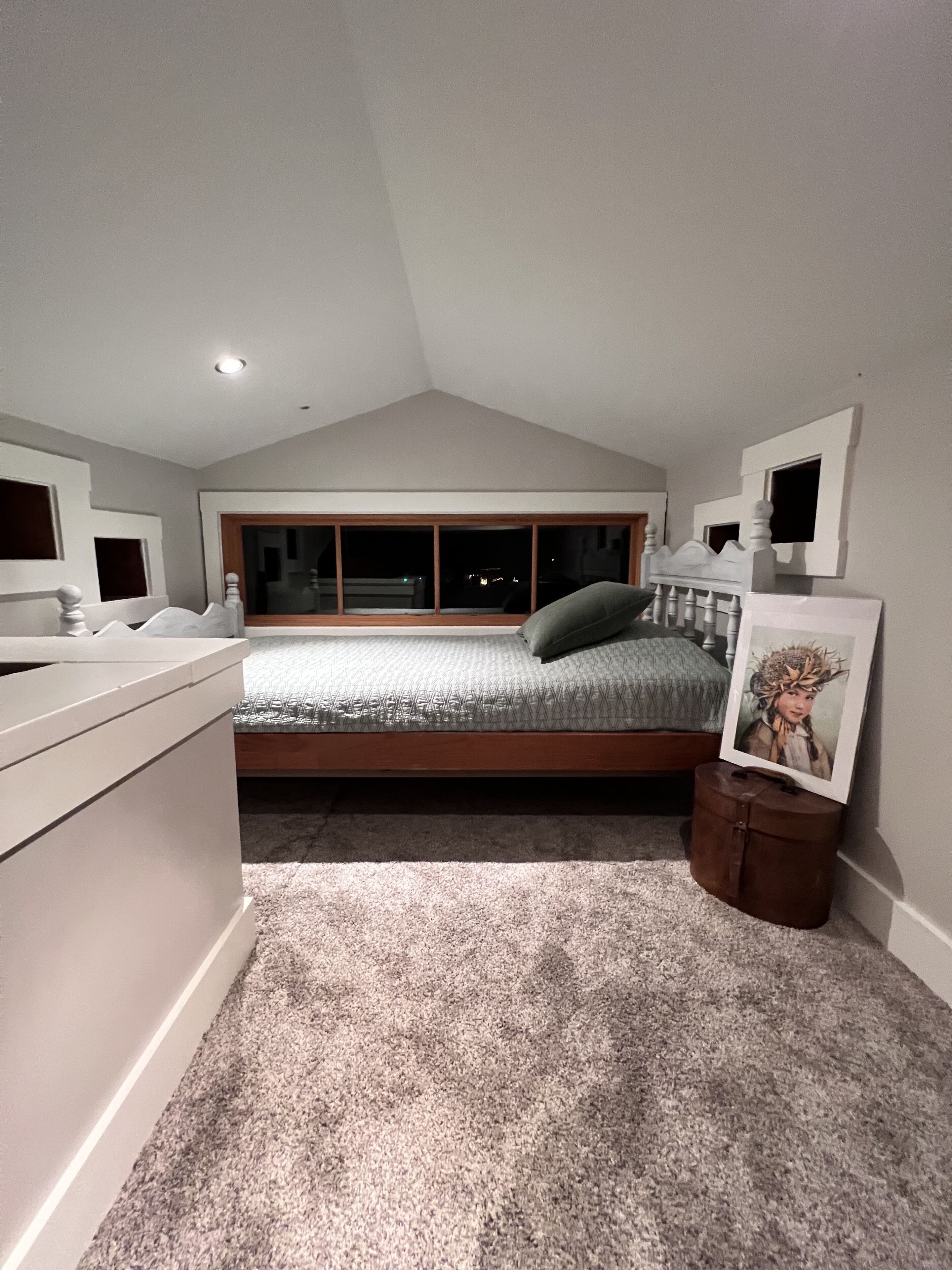 A bedroom with a sloped ceiling, a bed with a headboard, a nightstand, a framed picture, and a window with a view of the outside.