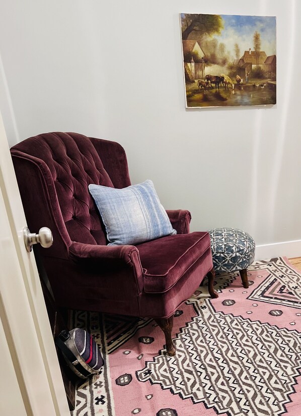 Une pièce comprenant un fauteuil en velours rouge foncé, un tapis à motifs et un tableau représentant un paysage rural accroché au mur.