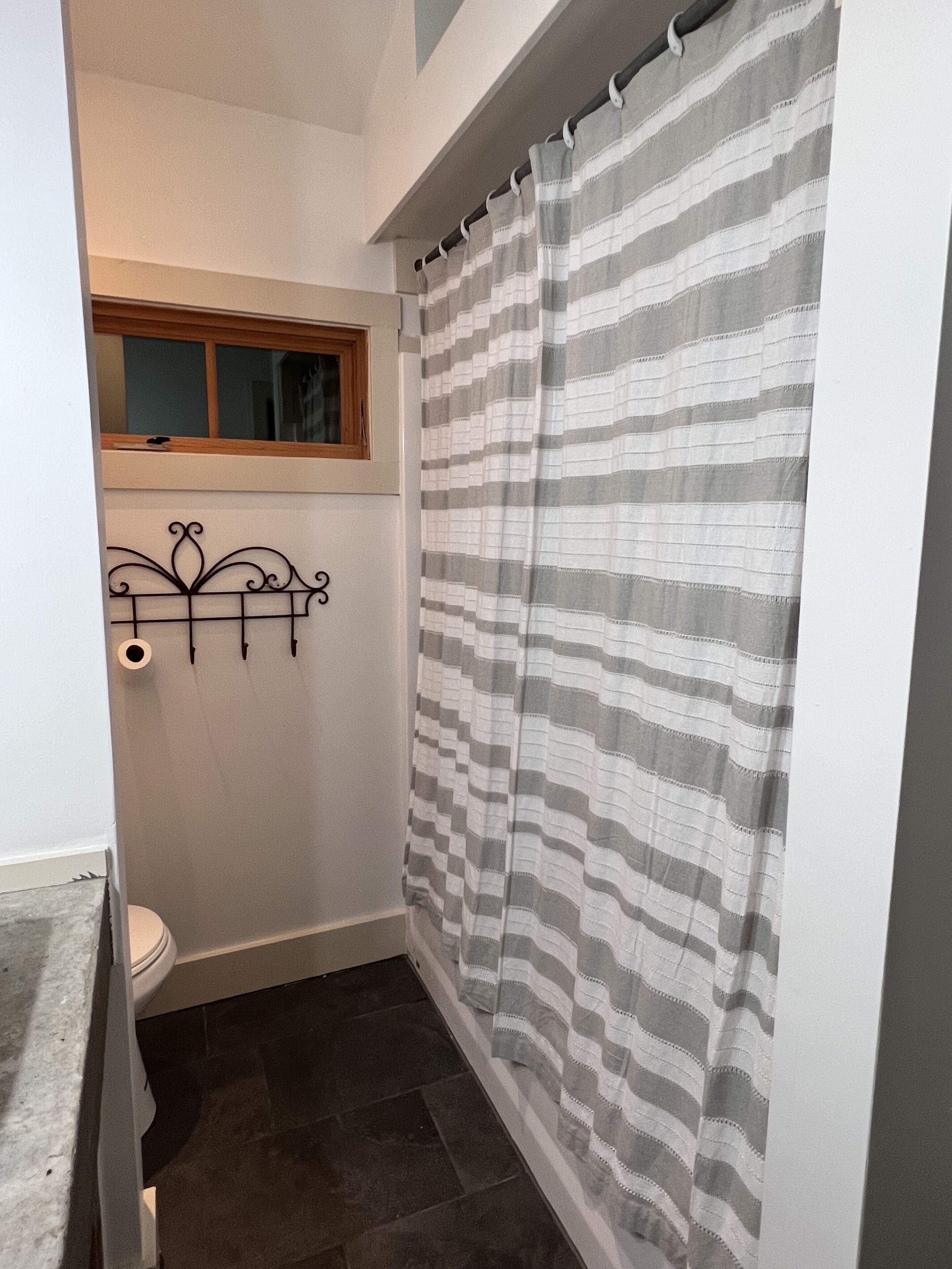A small, narrow bathroom with a shower curtain, a toilet, and a window above the toilet.