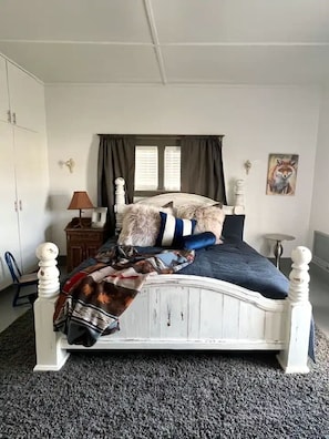 Ein Schlafzimmer mit einem weißen Himmelbett, einem Fenster mit Vorhängen, einer Lampe, einem Gemälde und einem Sessel.