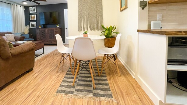 Una sala moderna con un sofá marrón, una mesa de comedor con sillas blancas, un ventilador de techo, un televisor montado en la pared y una zona de cocina con estantes de madera.