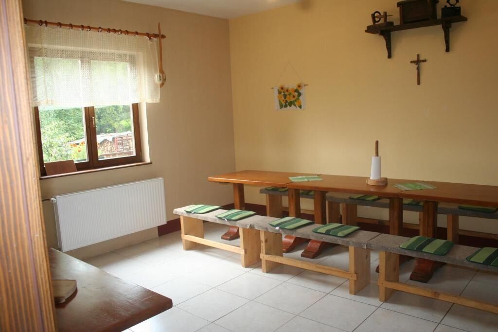 Una habitación con un largo banco de madera con cojines verdes, una ventana con cortina blanca, un radiador y una pared con una cruz y un adorno pequeño.