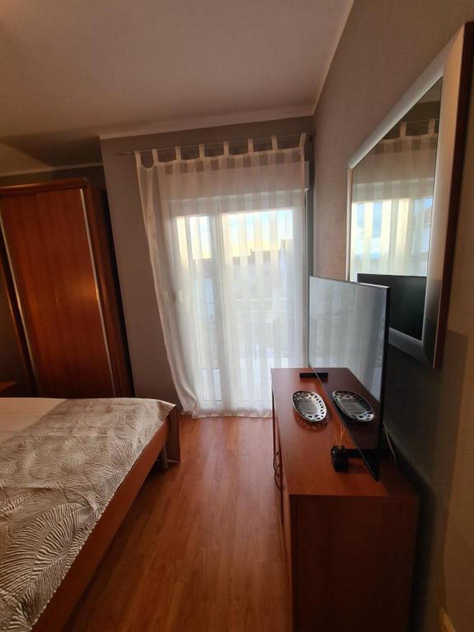 A hotel room with a bed, a desk with a computer, a television, and a window with sheer curtains.