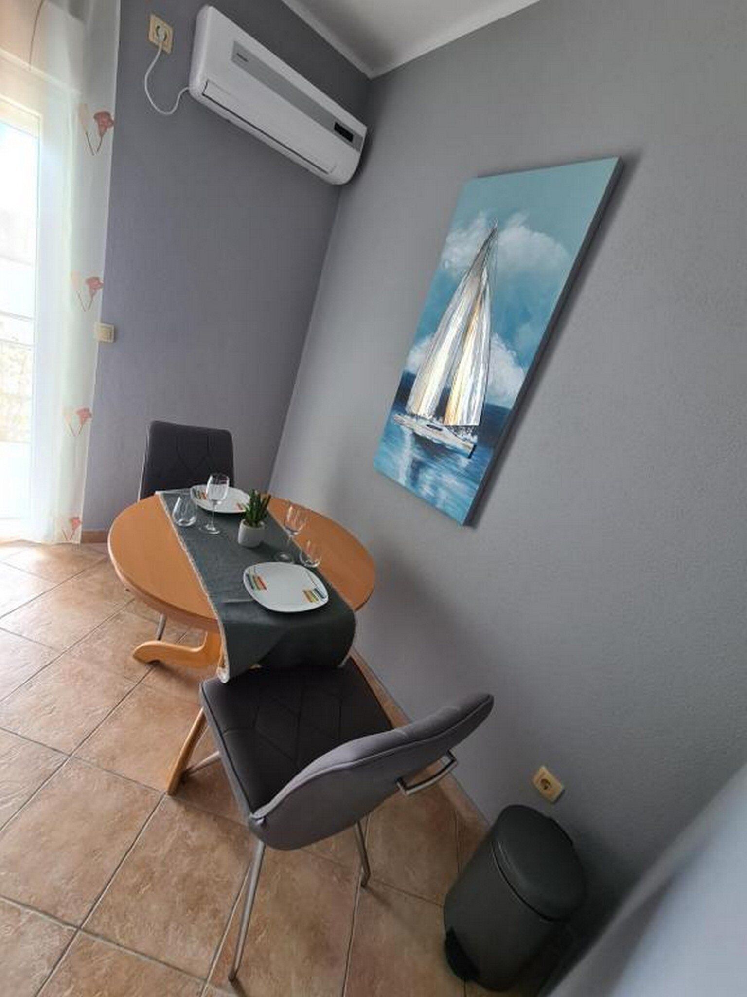 A dining area with a table set for two, a chair, a potted plant, and a painting of a sailboat on the wall.