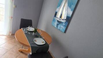 A dining area with a table set for two, a chair, a potted plant, and a painting of a sailboat on the wall.