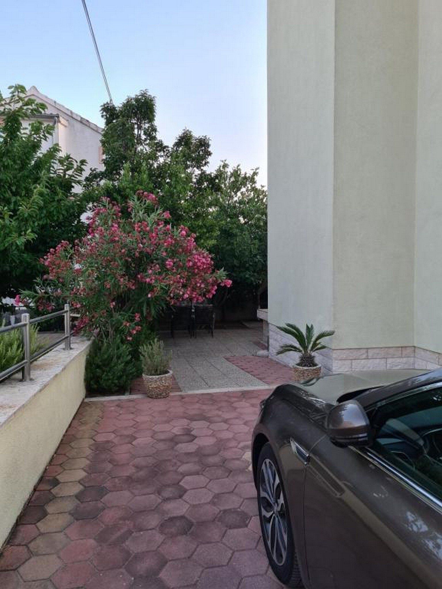 Een geplaveid pad dat leidt naar een auto, geparkeerd naast een gebouw met een grote struik roze bloemen.