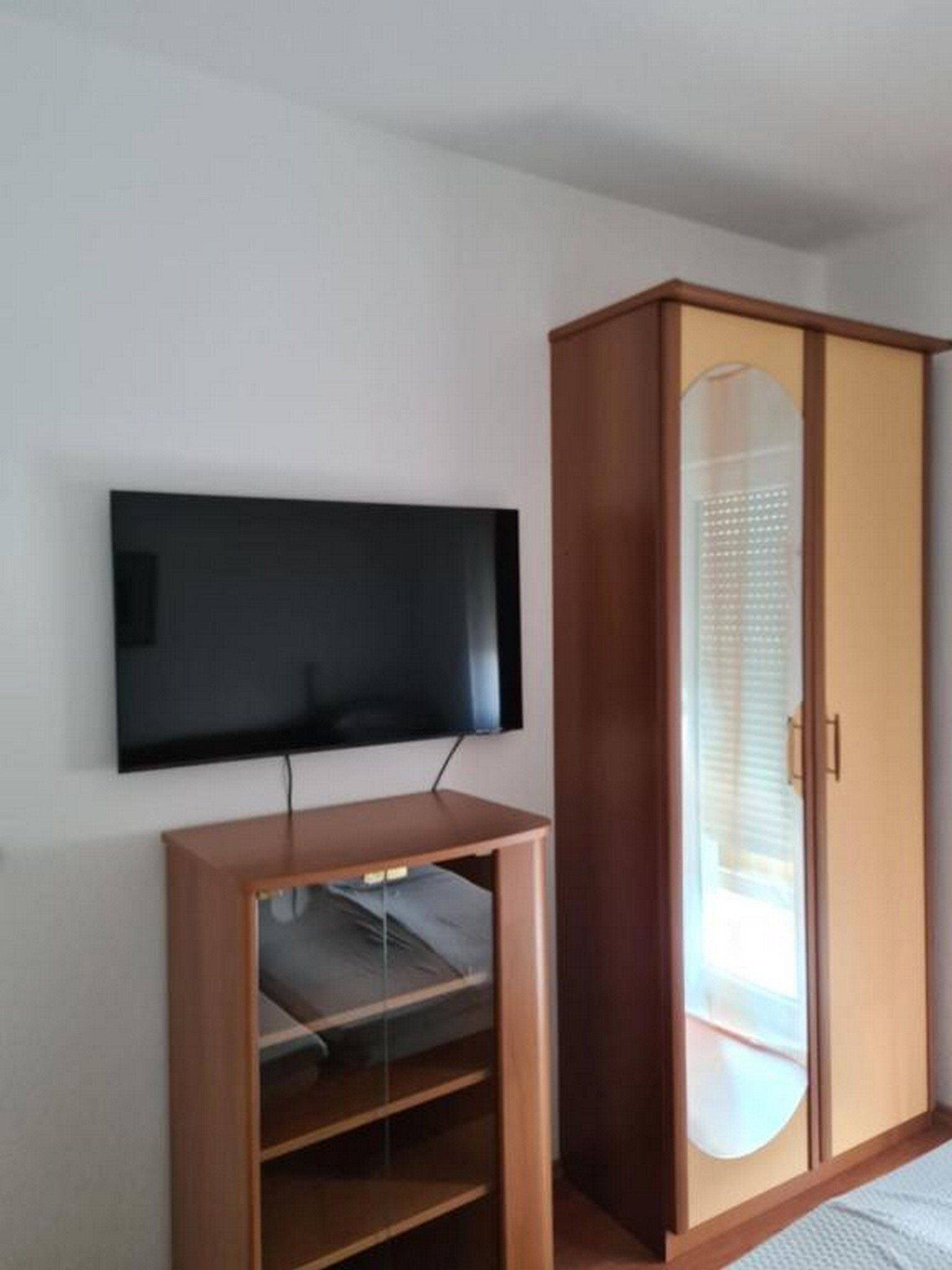 A room with a flat-screen TV mounted on the wall, a wooden wardrobe with a mirrored door, and a shelving unit below the TV.