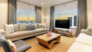 Ein modernes Wohnzimmer mit Sofa, Couchtisch und Fernseher, großen Fenstern mit Vorhängen und Blick auf den Sonnenuntergang.