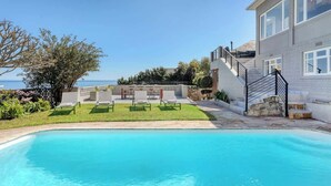 Ein Pool mit freiem Blick auf das Meer, eine Rasenfläche mit Liegestühlen und ein Gebäude mit einer Treppe.