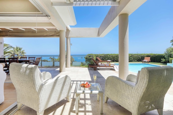 A covered outdoor seating area with wicker chairs, a glass table with fruit and a glass, and a view of the sea.