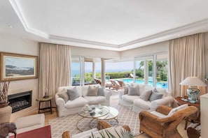 A spacious living room with a fireplace, two white sofas, a coffee table, and a view of a swimming pool.