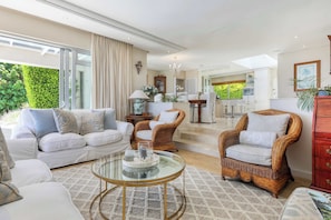 A spacious living room with a sofa, two wicker chairs, a glass coffee table, and a kitchen in the background.