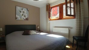Ein Schlafzimmer mit Bett, Nachttisch, Stuhl, Fenster mit Vorhängen und Heizkörper.