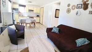 Ein Wohnzimmer mit einem dunkelbraunen Sofa, zwei Kissen, einem Couchtisch und einem Fernseher. Im Hintergrund befindet sich ein Essbereich mit Tisch und Stühlen.