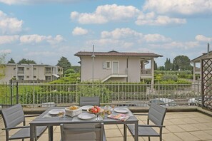 A terrace with a table set for dining, surrounded by chairs, with a view of residential buildings and greenery. - Appartamento Magenta (Moniga del Garda)