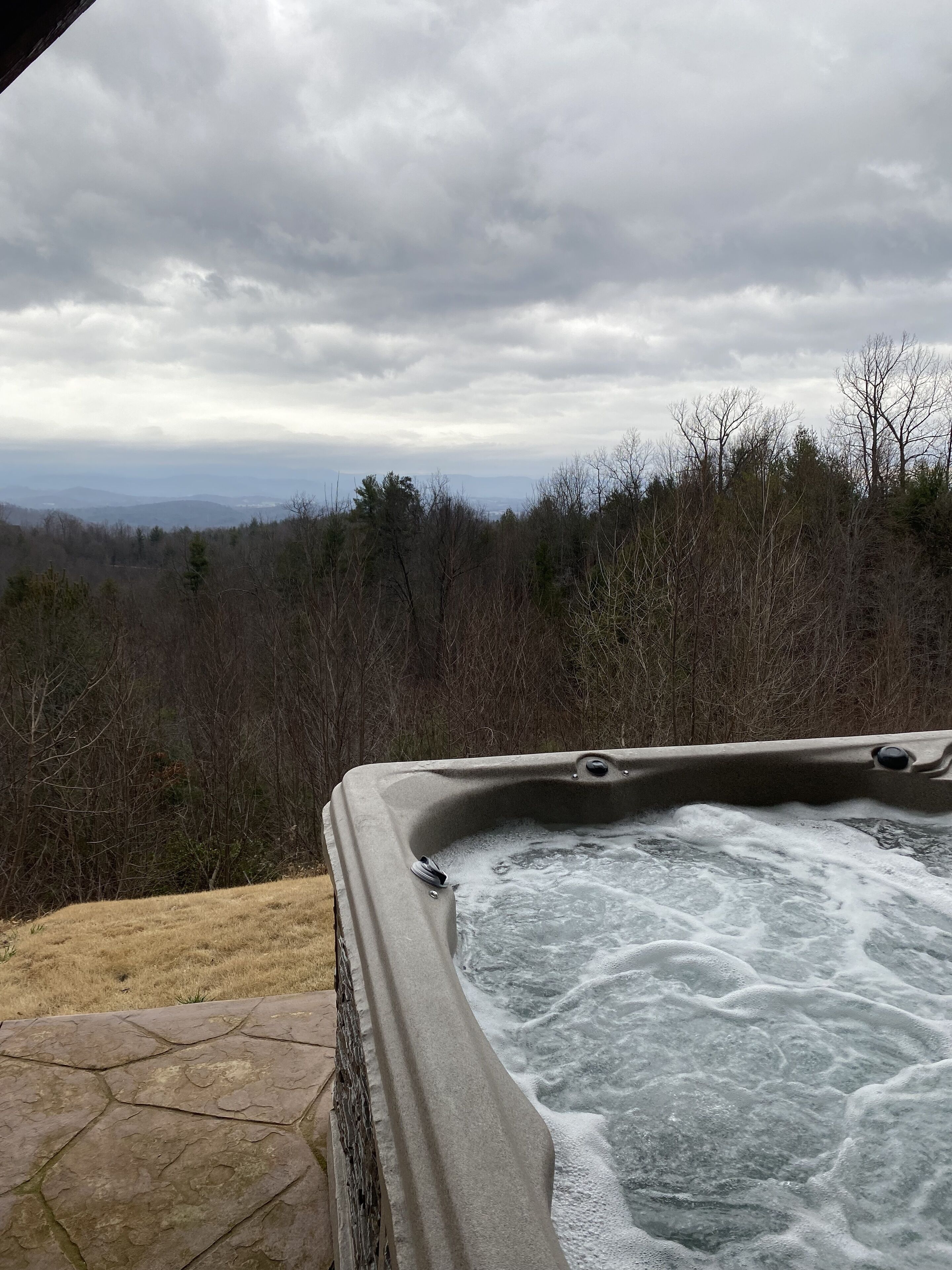 A hot tub with a stone patio, surrounded by a forested area and a view of distant mountains.