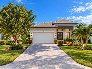 A two-story house with a paved driveway, a garage, and a well-maintained garden with various plants and a palm tree. - 779 Palm Paradise | Brand New Designer Pool Home (Naples)
