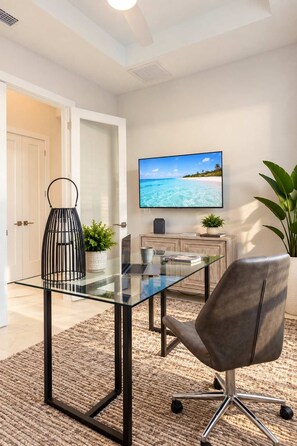 A modern office space with a glass table, leather chair, and a TV displaying a beach scene.