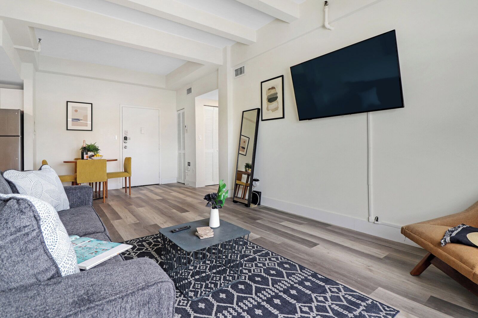 Ein modernes Wohnzimmer mit einem grauen Sofa, einem Couchtisch mit einer Vase, einem gemusterten Teppich, einem an der Wand montierten Fernseher und einem Essbereich mit einem gelben Stuhl.
