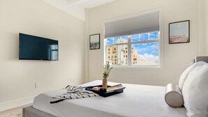 Et moderne hotelværelse med en stor seng, et fladskærms-tv monteret på væggen, et vindue med udsigt over byen og et lille bord med en vase og bøger.
