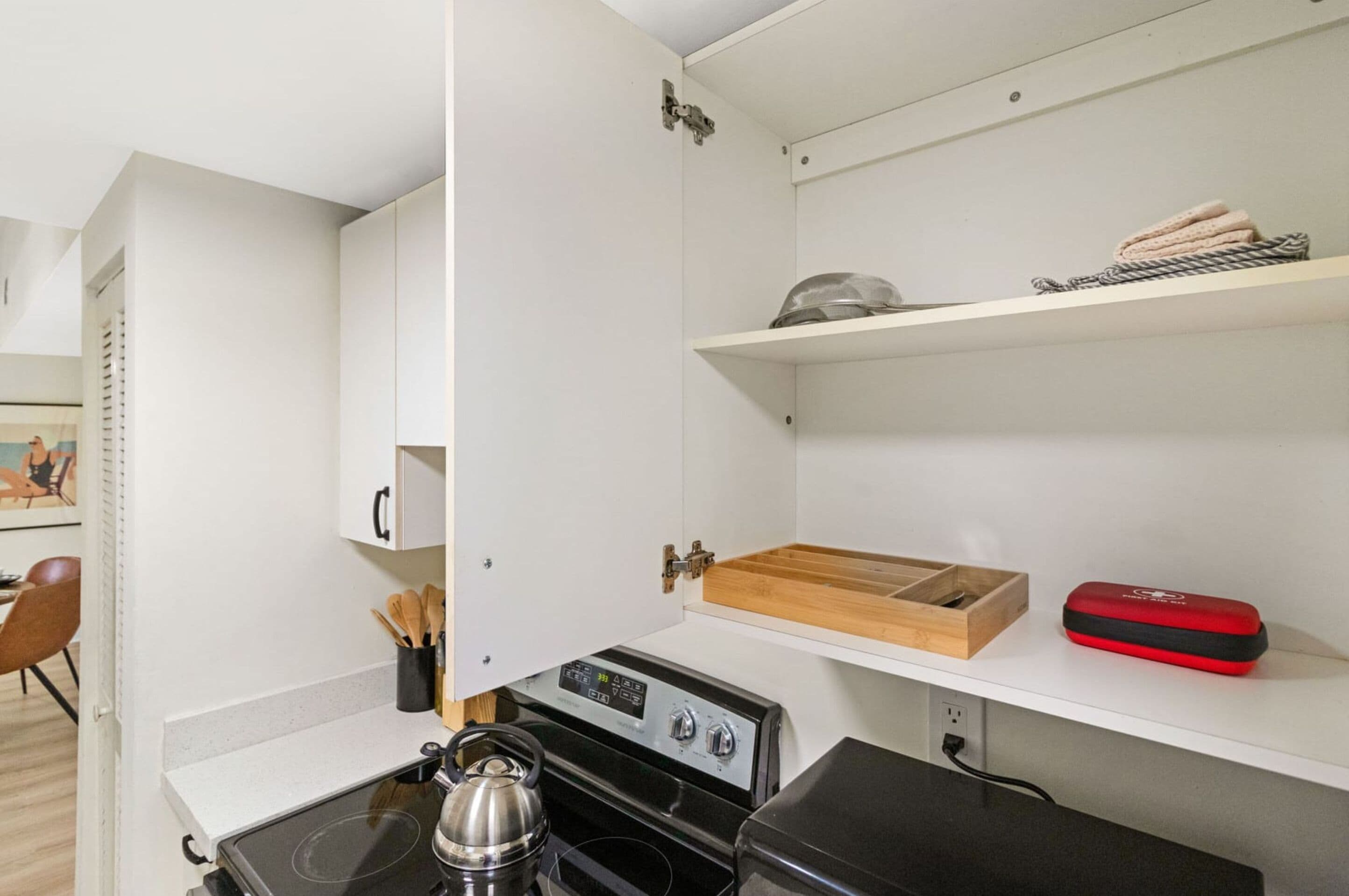 Une petite cuisine équipée d’une cuisinière, d’une bouilloire et d’un petit plateau en bois. On y trouve des placards blancs et une étagère avec des serviettes.