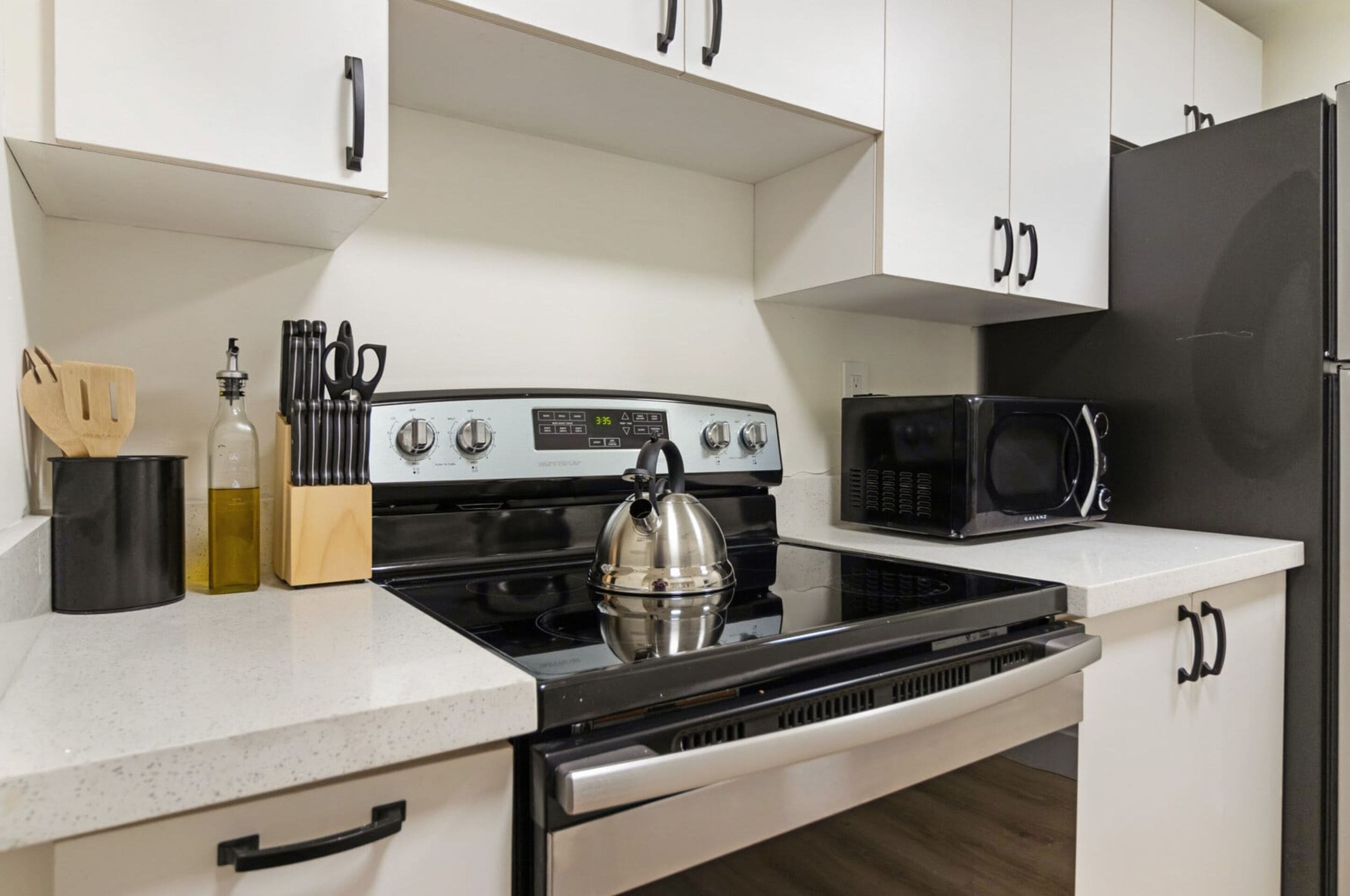 A modern kitchen with white cabinets, a stainless steel stove, a kettle, a microwave, and a set of knives.