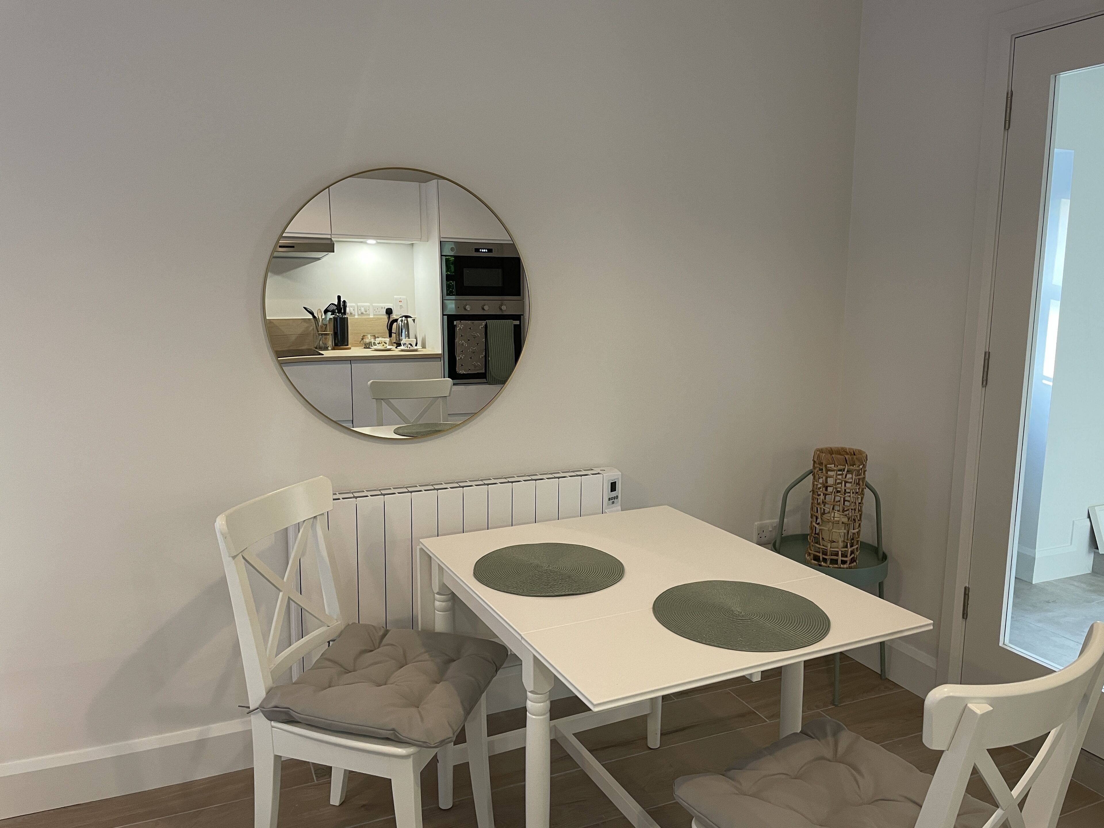 Un coin repas avec une table blanche, deux chaises, un miroir rond et une cuisine en arrière-plan.