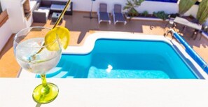 Un verre d’eau avec une tranche de citron, posé sur une table blanche, à côté d’une piscine où l’on aperçoit des chaises longues au fond.