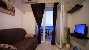 A room with a sofa, a television on a shelf, a potted plant, and a view of the sea through a glass door. - Modern vacation apartment with pool and sea view (Pisak)