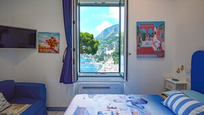 Ein Schlafzimmer mit einem Bett, einem blauen Sofa, einem Fenster mit Blick auf eine Küstenlandschaft und zwei Wandbildern.