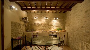 Una cocina con paredes de piedra, una mesa redonda de cristal y sillas de metal, un pequeño estante con utensilios de cocina y un techo de madera.
