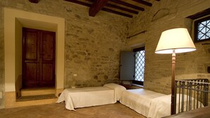Una habitación rústica con dos camas individuales, una puerta de madera, paredes de piedra, una ventana con rejas y una lámpara de pie.