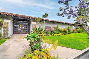 A historical museum with a tiled roof, a concrete pathway leading to the entrance, and a well-maintained garden with various plants and a tree with purple flowers.