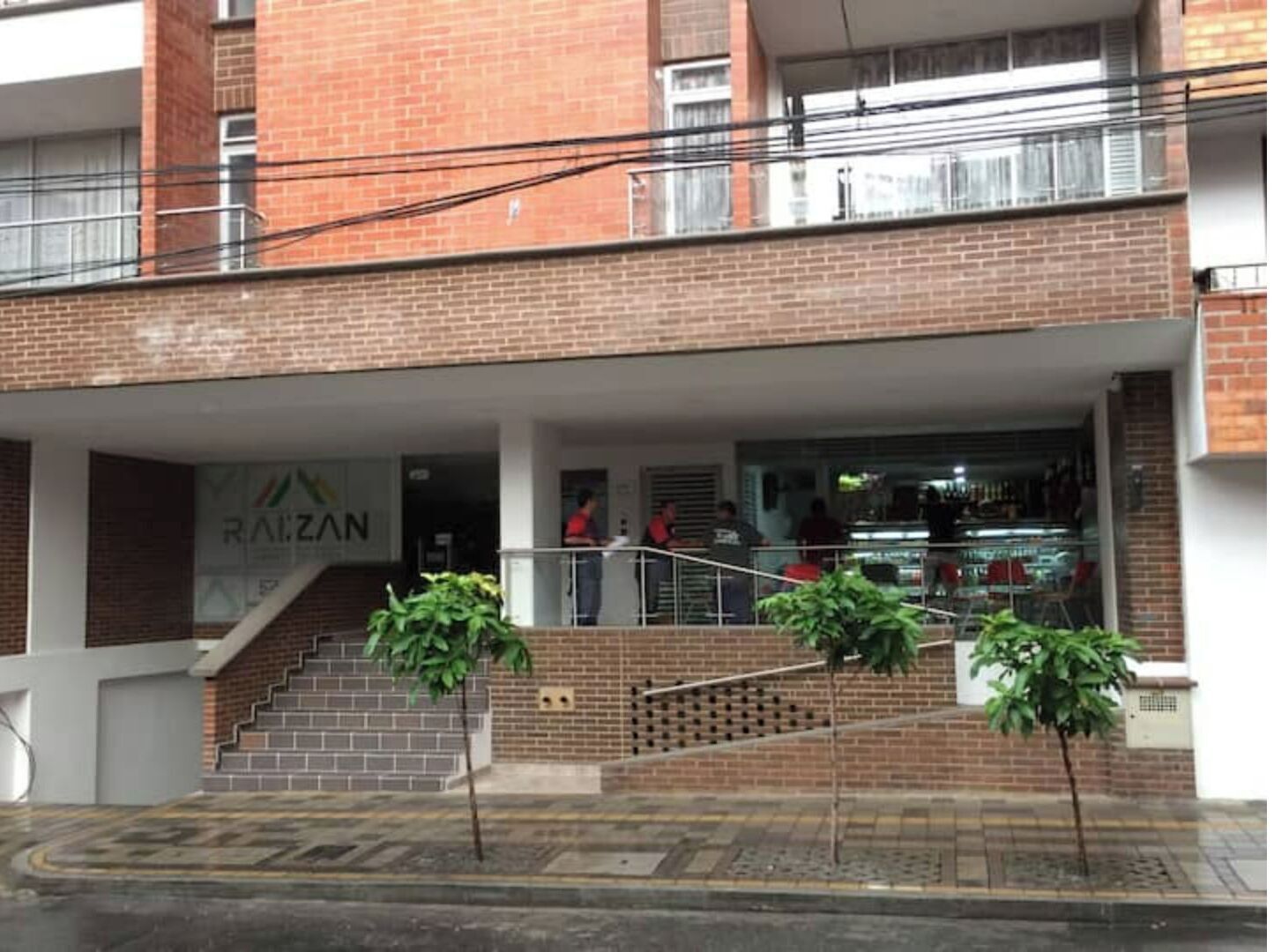 A multi-story building with a brick facade, a covered entrance area, and a visible storefront with a sign reading "RAIZAN".
