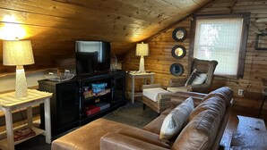 Une pièce mansardée chaleureuse avec un plafond en bois, un téléviseur posé sur un meuble, un canapé et deux fauteuils.