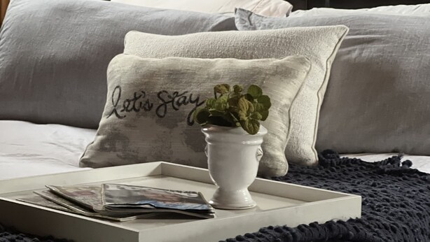 Un lit bien fait, avec un oreiller blanc orné du texte « Restons ensemble » et une petite plante en pot.