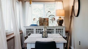 Un coin repas avec une table blanche, des chaises noires, un ventilateur de plafond, une fenêtre avec des rideaux blancs, une lampe et une horloge au mur.
