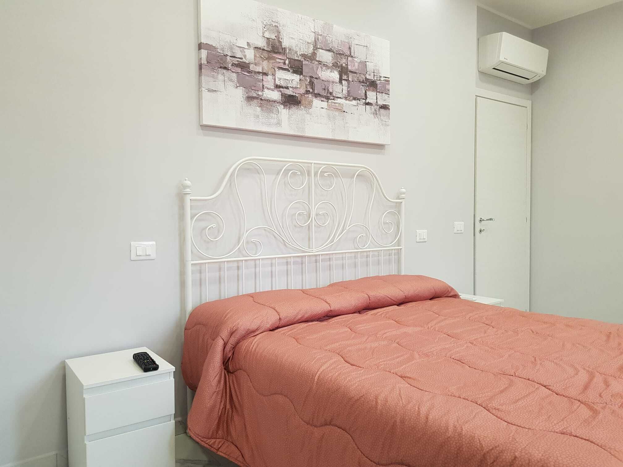 Un dormitorio con cabecera de metal blanco, colcha rosa, mesita de noche blanca con un control remoto y un aire acondicionado instalado en la pared.