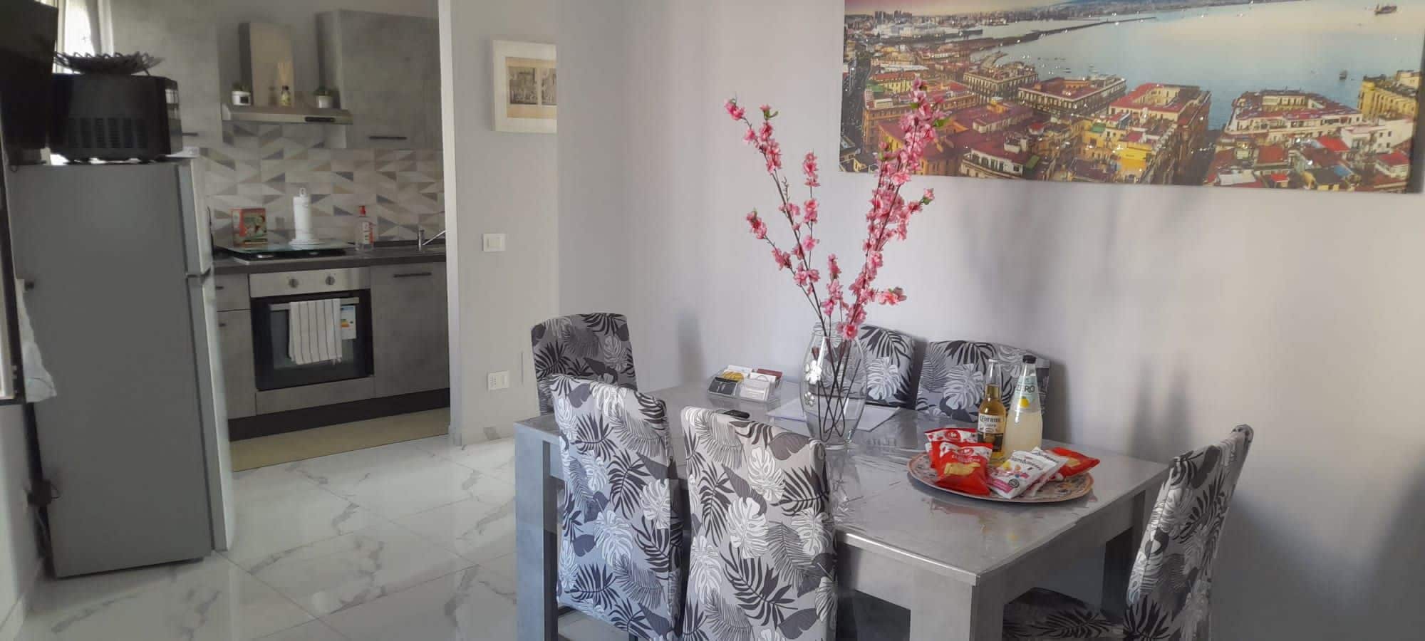 Un comedor con una mesa para dos, sillas con estampado de hojas, un florero con flores rosadas y un cuadro de un paisaje urbano en la pared.
