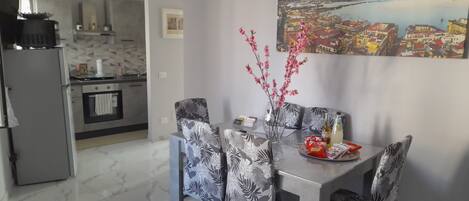 Un comedor con una mesa para dos, sillas con estampado de hojas, un florero con flores rosadas y un cuadro de un paisaje urbano en la pared.