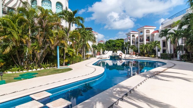 A resort pool area with a curved pool, palm trees, and a white building with arched windows. - Luxury 3 Bdrm Condo in Playacar. Tennis & Golf Close By, Shops Walking Distance! (Playa del Carmen)