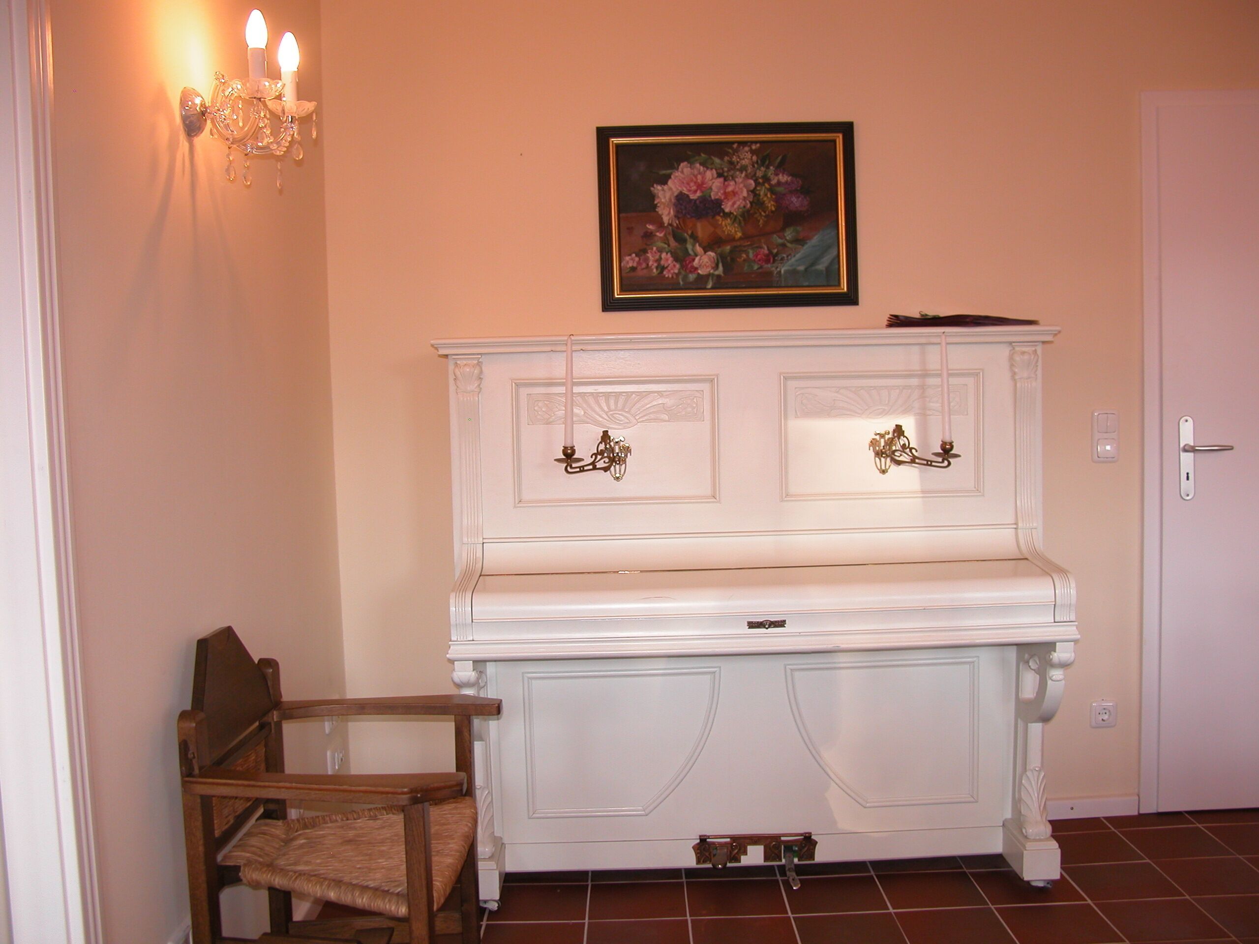 Ett vitt piano med utsmyckade detaljer, en trästol, en tavla som hänger på väggen och en vägglampa.