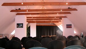 Ein Zimmer mit schrägen Wänden, Holzbalken und einer Chandelüster. Stühle sind in Reihen vor einer Bühne angeordnet.